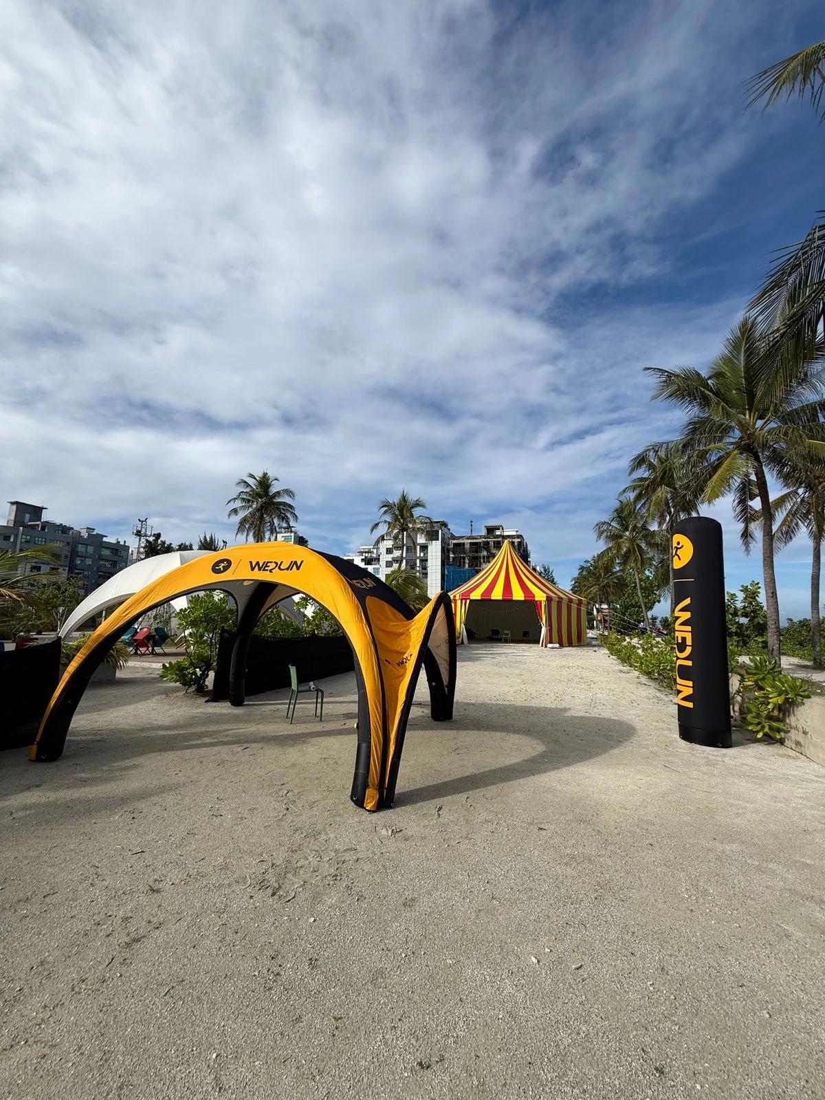 A wide outdoor shot of a beach event site, showcasing a yellow and black inflatable arch tent branded "WEQUN," a matching inflatable pillar, and a red and yellow striped circus-style tent, situated on sandy ground with palm trees under a blue, cloudy sky.